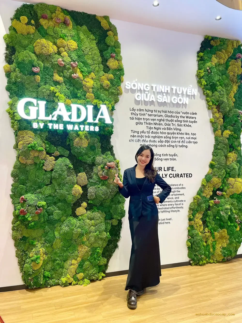 Ms Hòa đứng trước bức tường rêu logo Gladia By The Waters tại sales gallery dự án